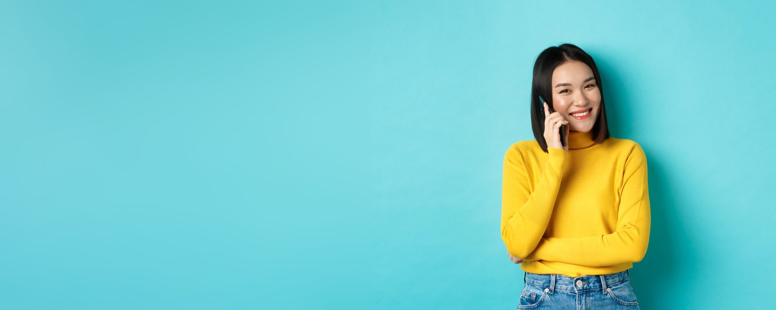 young stylish asian woman talking on phone, calling friend and smiling, standing with smartphone against blue background